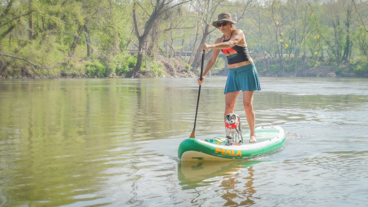 Paddler and her dog on a Hala inflatable paddleboard on a calm tree-lined river